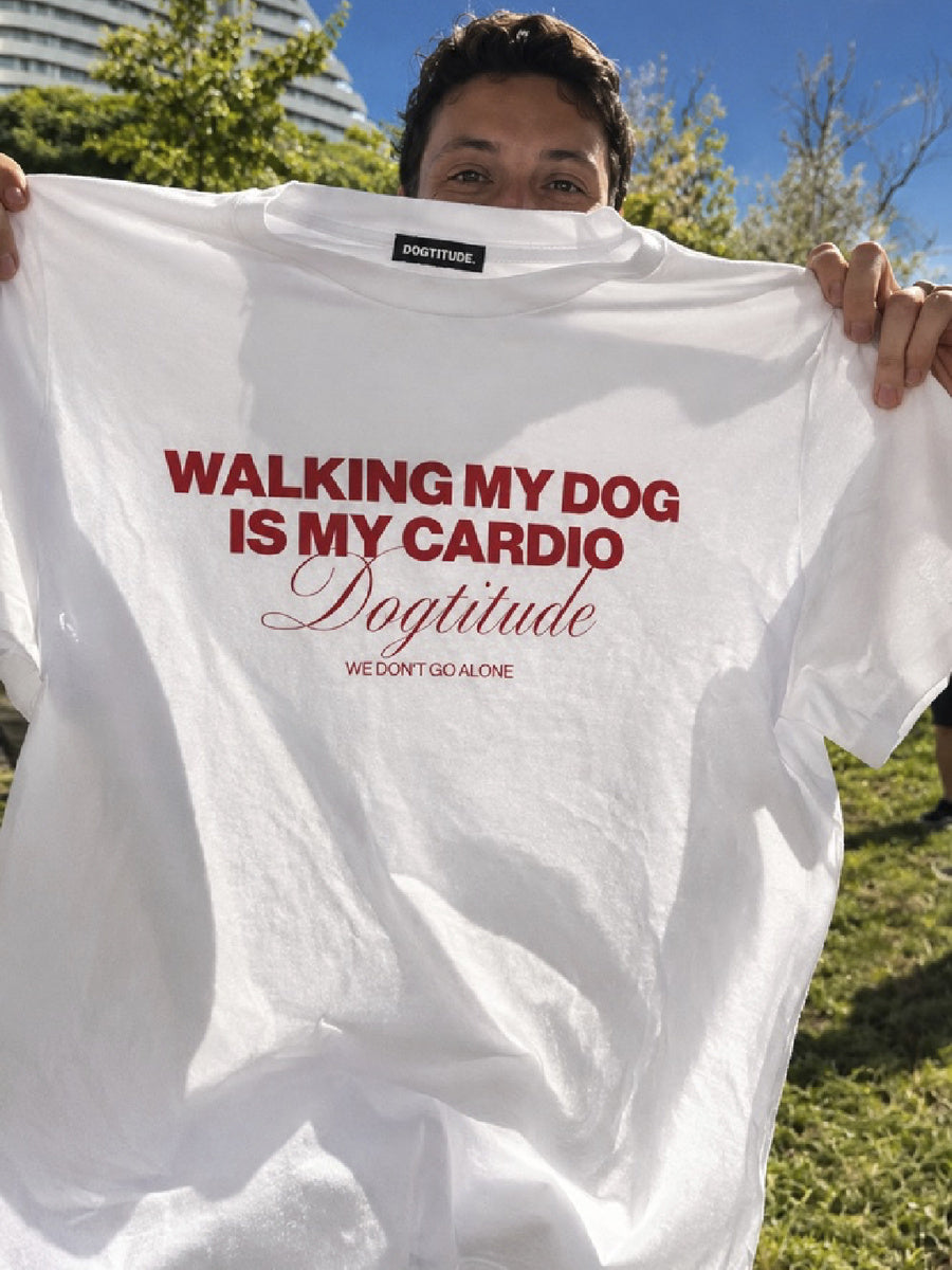 Detalle de una persona sosteniendo al aire libre la camiseta blanca DOGTITUDE con el estampado rojo WALKING MY DOG IS MY CARDIO.