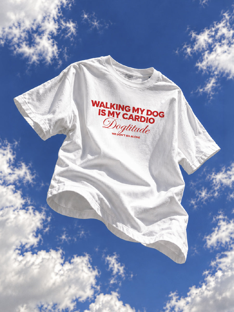 Camiseta blanca DOGTITUDE con estampado rojo WALKING MY DOG IS MY CARDIO sobre un cielo azul con nubes.